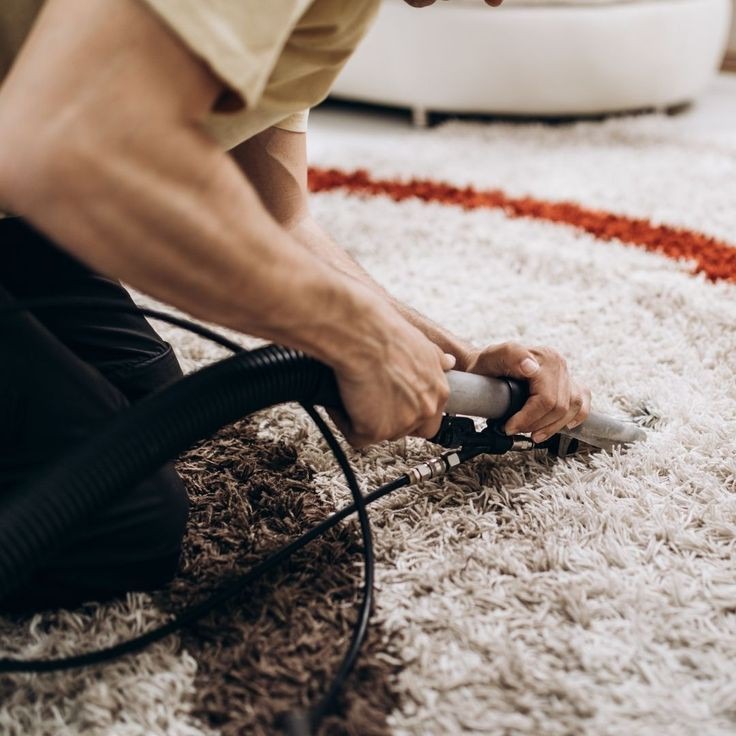 Czyszczenie jasnego dywanu z pomarańczowym pasem za pomocą odkurzacza piorącego z czarnym wężem.
