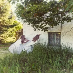 Dwie dziewczynki w białych sukienkach i skrzydłach anioła dotykają ściany starego budynku z zielonymi okiennicami, otoczonego wysoką trawą i drzewami.