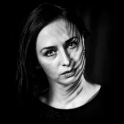 Portret kobiety w czerni i bieli z cieniem przypominającym liście na twarzy, oświetlonej, z błyszczącymi ustami i intensywnym spojrzeniem.