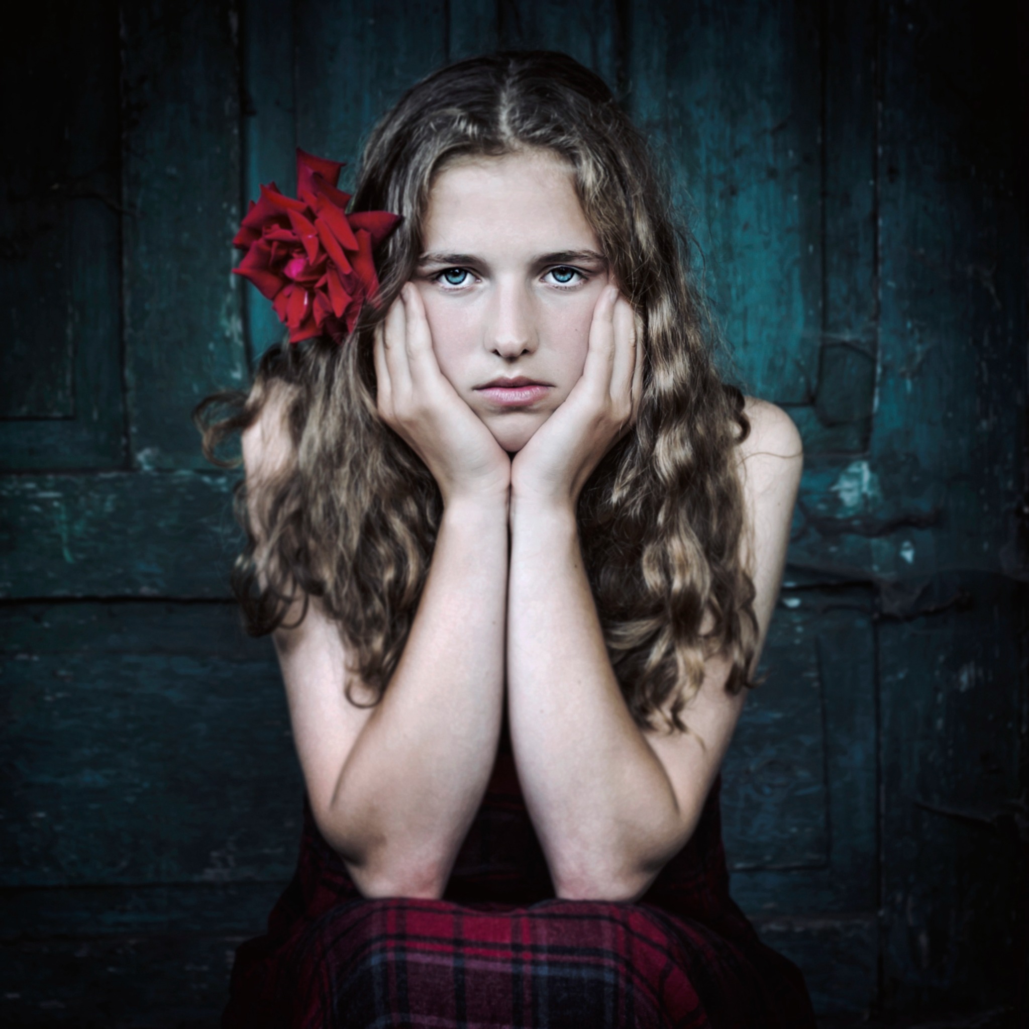 Portret młodej dziewczyny o jasnych oczach z czerwoną różą we włosach, opierającej brodę na dłoniach, na tle turkusowych, drewnianych drzwi.