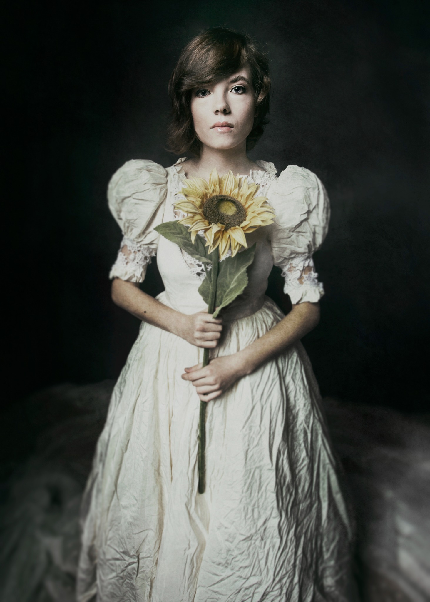 Młoda kobieta w kremowej sukni z bufiastymi rękawami i koronkowymi wstawkami, trzymająca pojedynczego słonecznika na ciemnym tle.