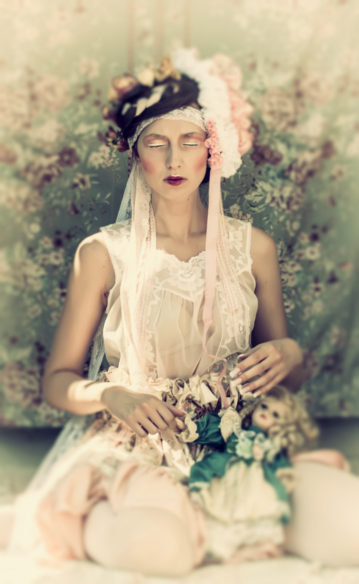 Kobieta z zamkniętymi oczami w stylizowanej sukience i nakryciu głowy z kwiatów, trzymająca lalkę na tle wzorzystej tapety.