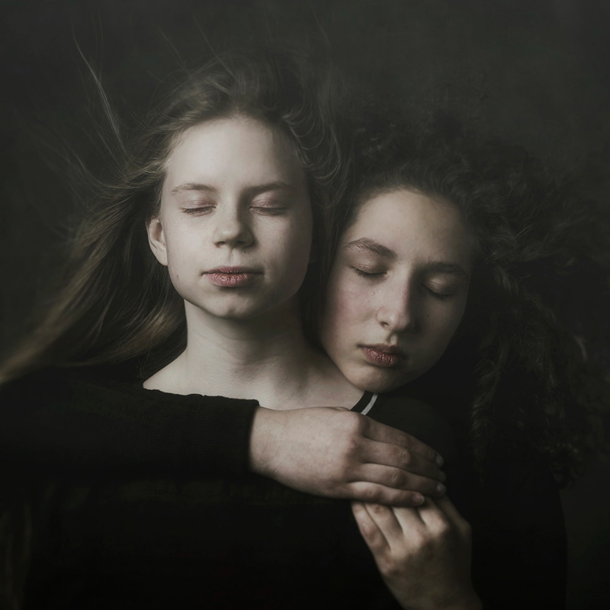 Portret dwóch nastolatek z zamkniętymi oczami w ciemnym otoczeniu, jedna obejmuje drugą, włosy rozwiane, subtelne światło.