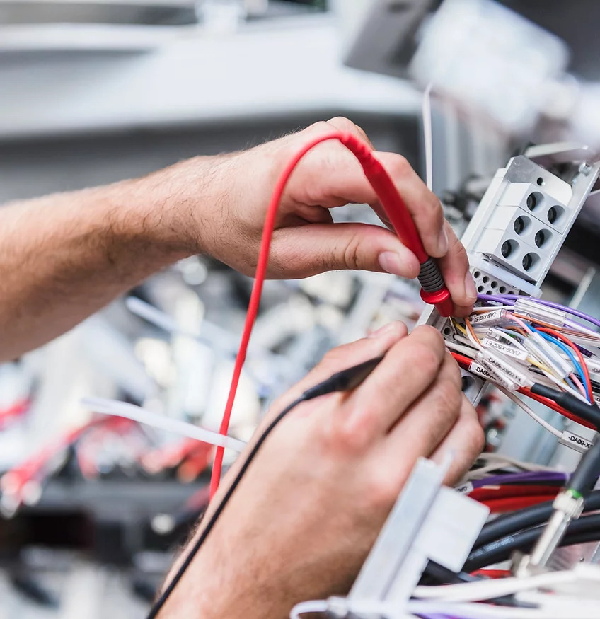 Elektryk mierzy napięcie w rozdzielnicy za pomocą miernika z czerwonym przewodem, widoczne kolorowe kable z etykietami.