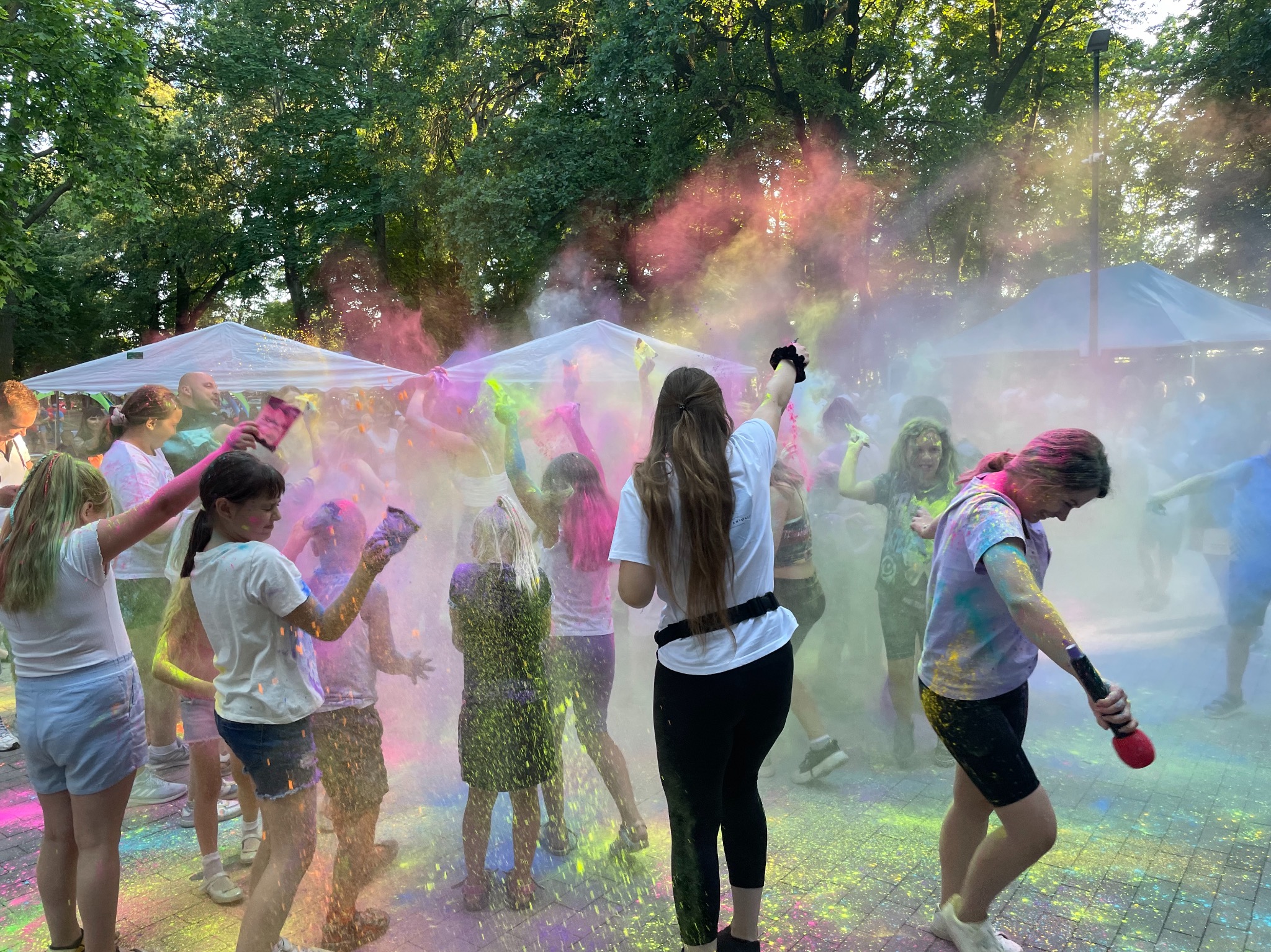Grupa dzieci i dorosłych rzuca kolorowymi proszkami Holi podczas imprezy plenerowej pod białymi namiotami, z drzewami w tle, w atmosferze zabawy i radości.