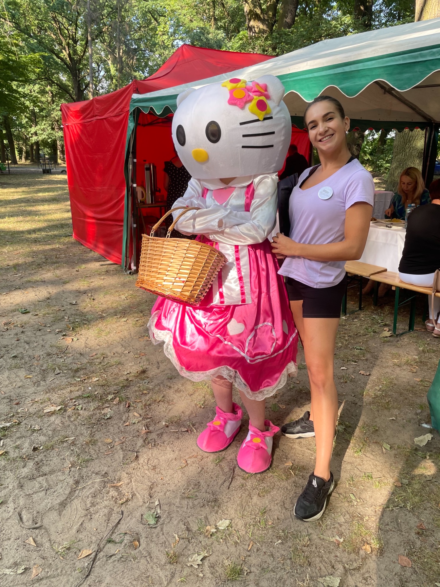 Animator w stroju Hello Kitty z koszykiem, obok uśmiechnięta kobieta w fioletowej koszulce, na tle czerwonego namiotu i zieleni parku.