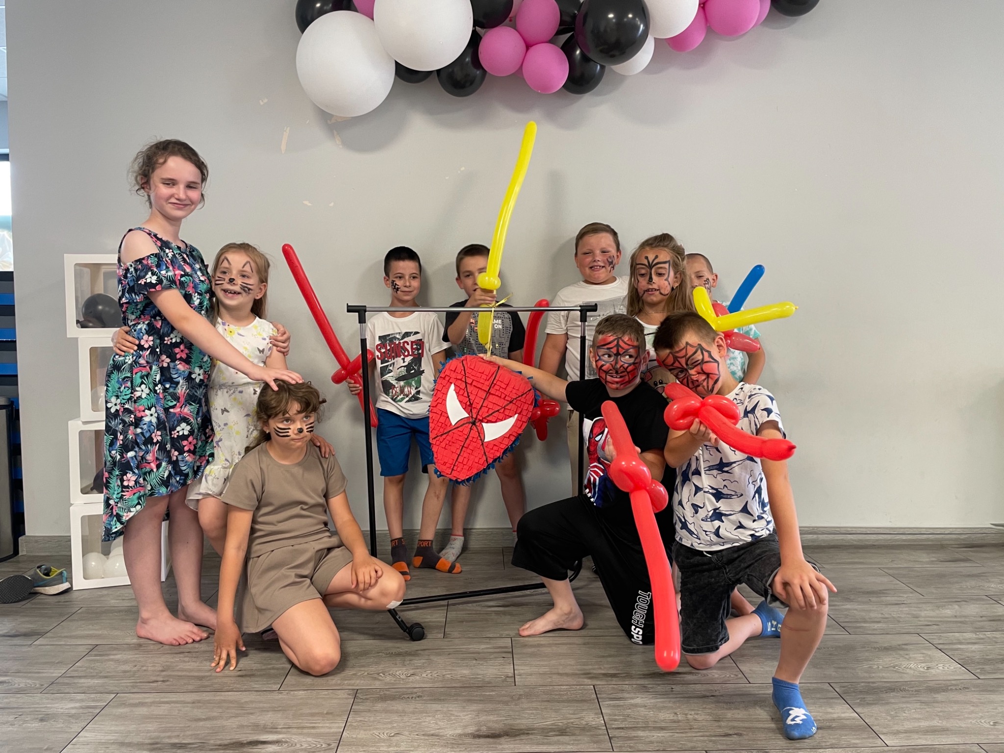 Grupa dzieci z pomalowanymi twarzami pozuje z balonami w kształcie mieczy i tarczą Spidermana na tle dekoracji z balonów w kolorach białym, różowym i czarnym.