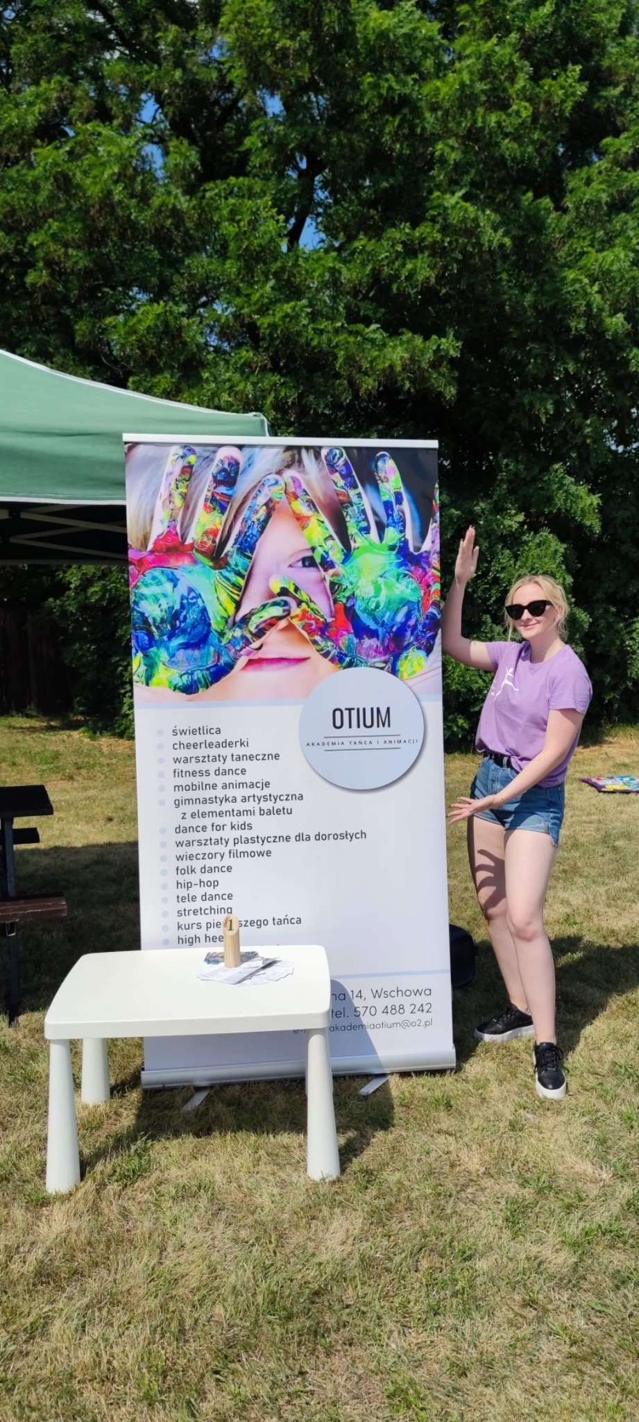 Kobieta w okularach przeciwsłonecznych pozuje obok roll-upu z ofertą akademii tańca i animacji na trawniku, widoczny stół z ulotkami.