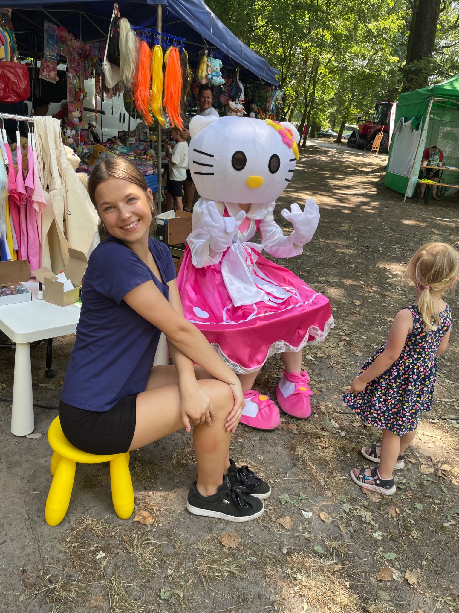 Uśmiechnięta kobieta siedzi na żółtym stołku obok osoby w kostiumie Hello Kitty, z zaciekawioną dziewczynką w sukience w serduszka i straganami w tle.
