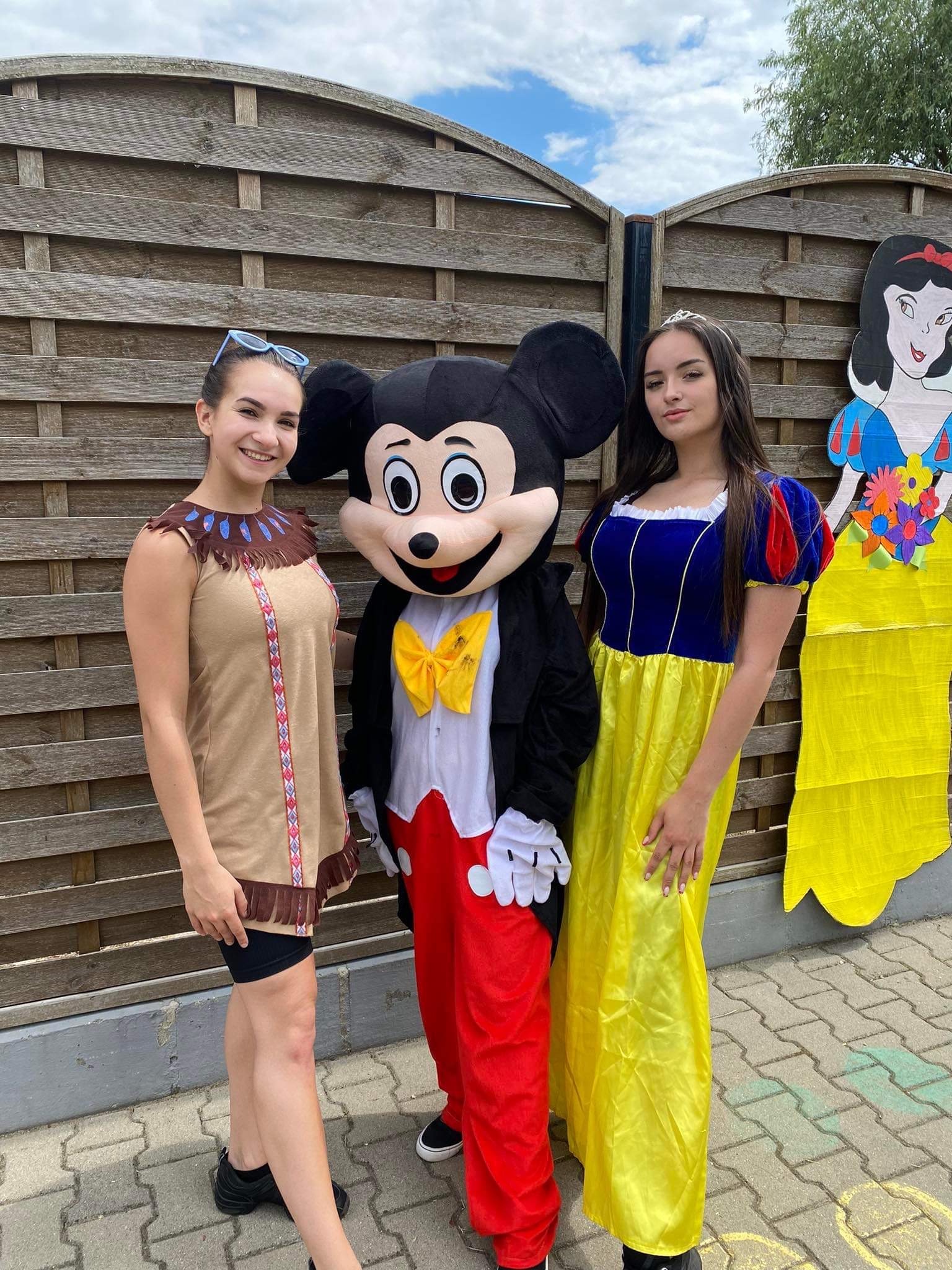 Dwie uśmiechnięte kobiety w kostiumach: jedna w stroju indiańskim z okularami na głowie, druga jako Królewna Śnieżka obok postaci w przebraniu Myszki Miki na tle drewnianego płotu i kartonowej...