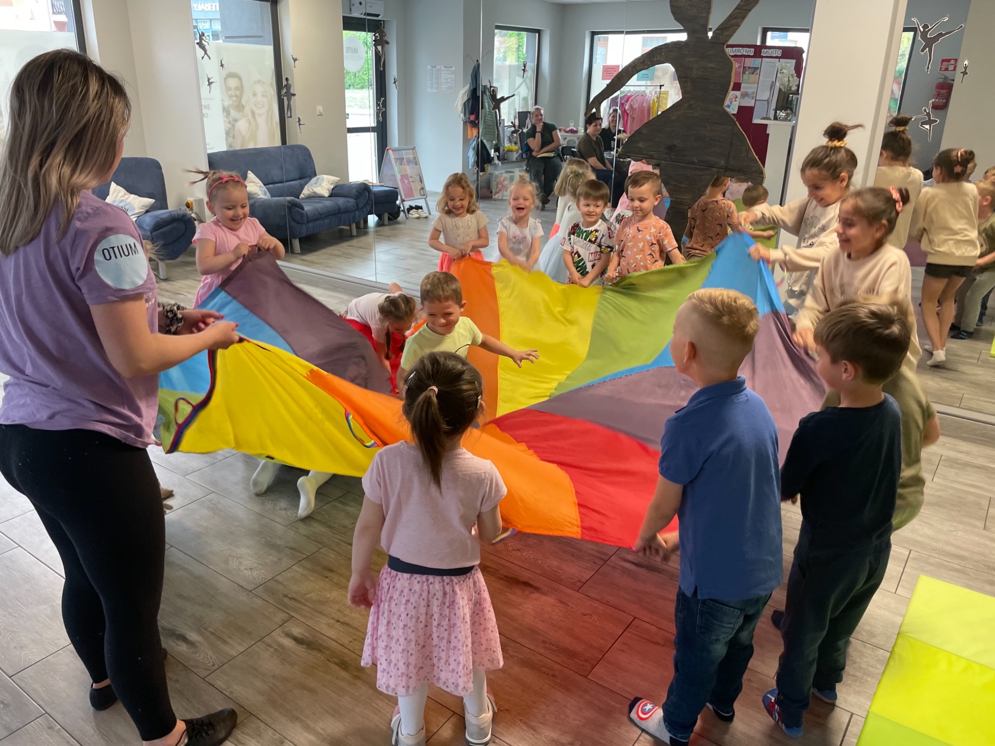 Grupa roześmianych dzieci z animatorem bawią się kolorową chustą animacyjną w przestronnym, jasnym pomieszczeniu z lustrami i drewnianą figurą baletnicy.
