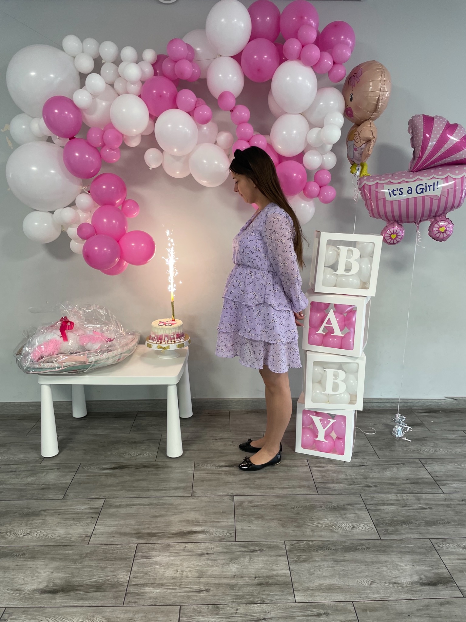Uroczystość baby shower: kobieta w kwiecistej sukience stoi obok stolika z tortem z fontanną iskier, dekoracje z białych i różowych balonów, napisy BABY w ozdobnych pudełkach i balon w kształcie...