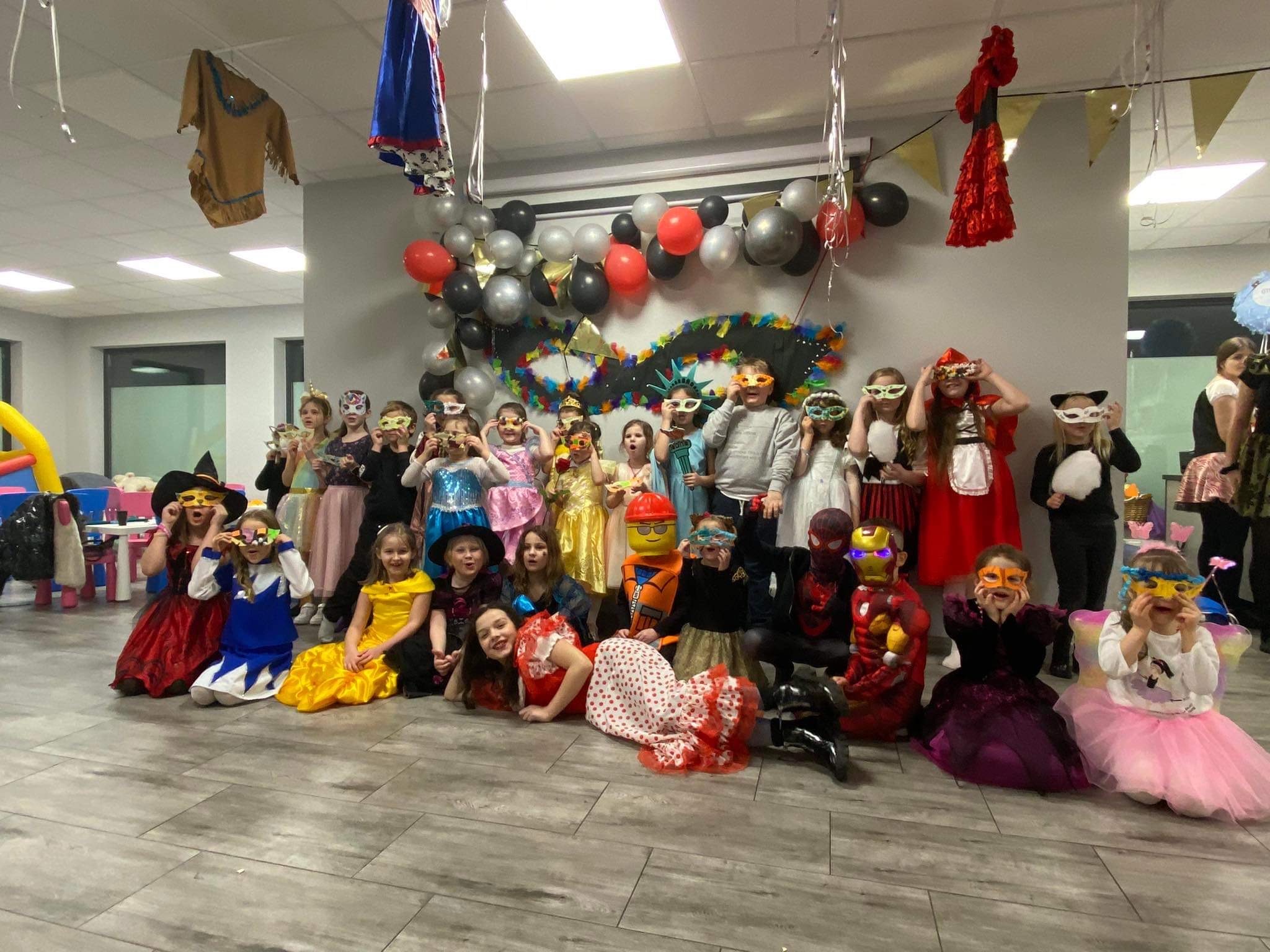 Duża grupa dzieci w różnorodnych kostiumach i maskach pozuje do zdjęcia na tle szarej ściany udekorowanej balonami i dekoracjami karnawałowymi.