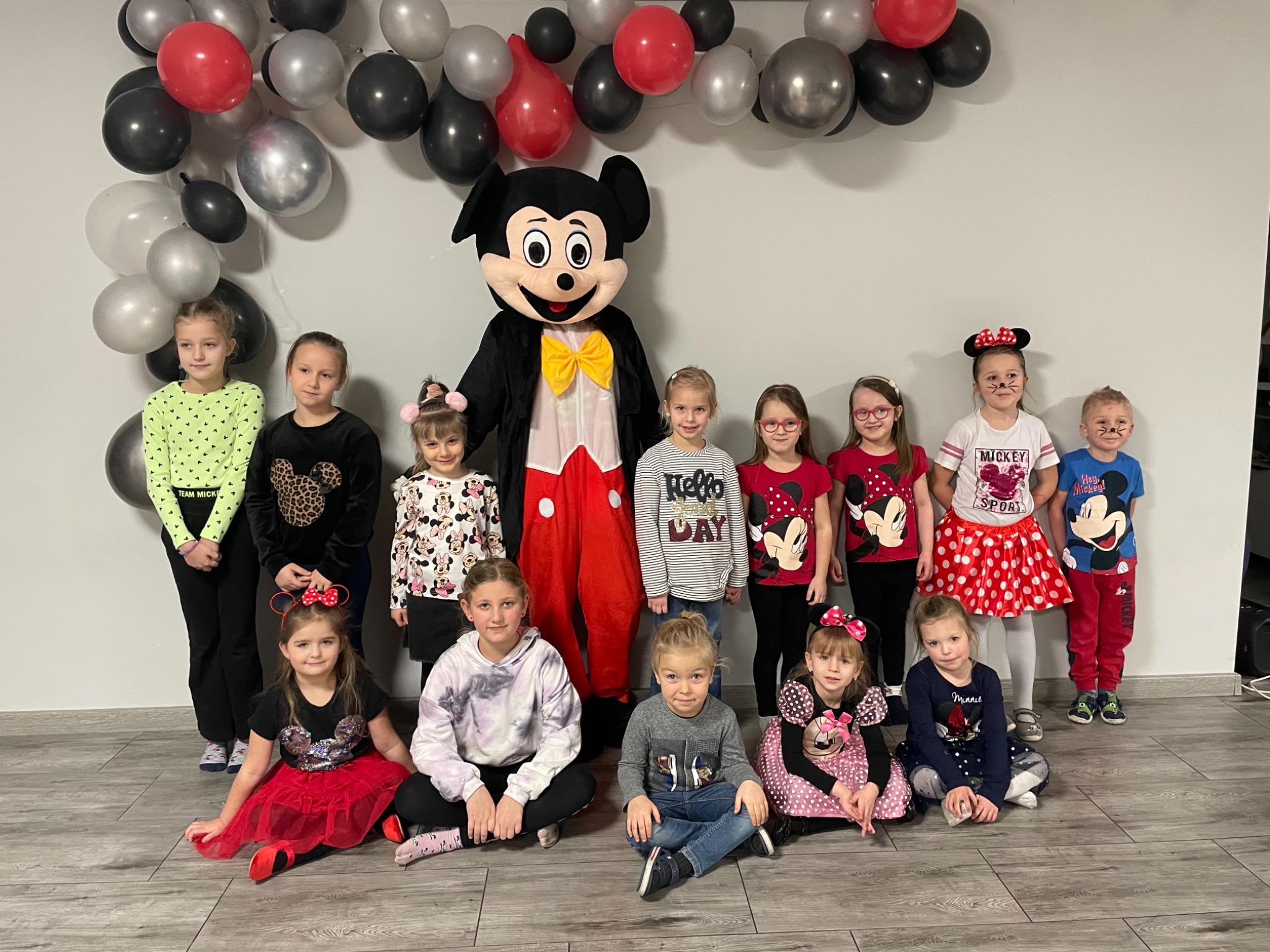 Grupa dzieci pozuje do zdjęcia z osobą w kostiumie Myszki Miki, za nimi dekoracja z czarnych, srebrnych i czerwonych balonów.