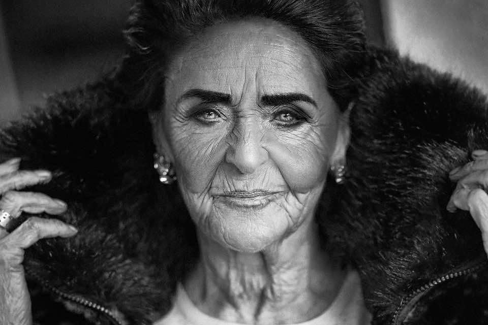 Czarno-biały portret starszej kobiety z wyrazistymi zmarszczkami, ubranej w futrzaną kurtkę, z kolczykami w uszach i pierścionkiem na palcu.