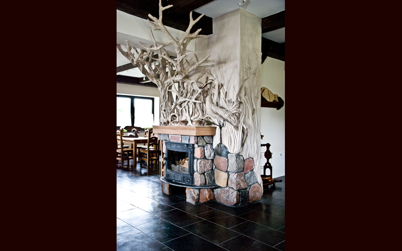 Kominek obudowany kamieniami i dekoracjami rzeźbiarskimi, z abstrakcyjną rzeźbą z gałęzi powyżej i płaskorzeźbą z motywami roślinnymi na kolumnie, w tle widoczne stoły i krzesła
