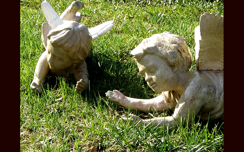 Dwie rzeźby aniołków z betonu lub kamienia leżące w trawie, jeden z uszkodzonym skrzydłem, skierowane ku sobie.