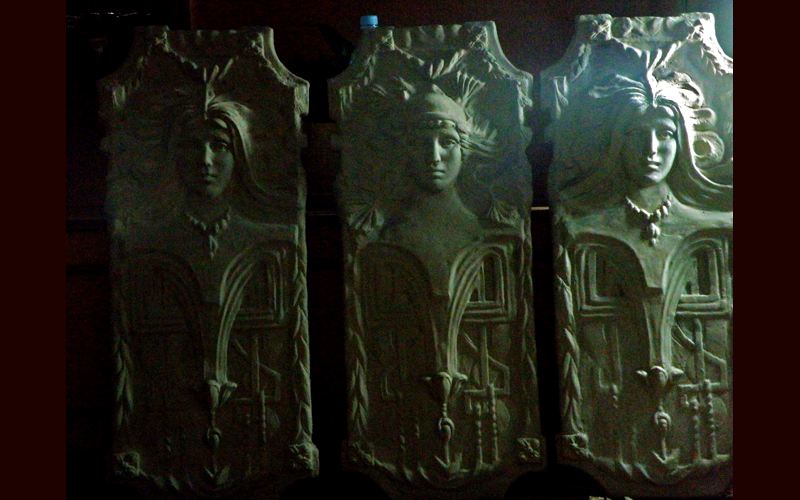 Trzy pionowe rzeźbione panele z wizerunkami kobiecych postaci w stylu art deco, oświetlone z góry, z widocznymi detalami ornamentów.