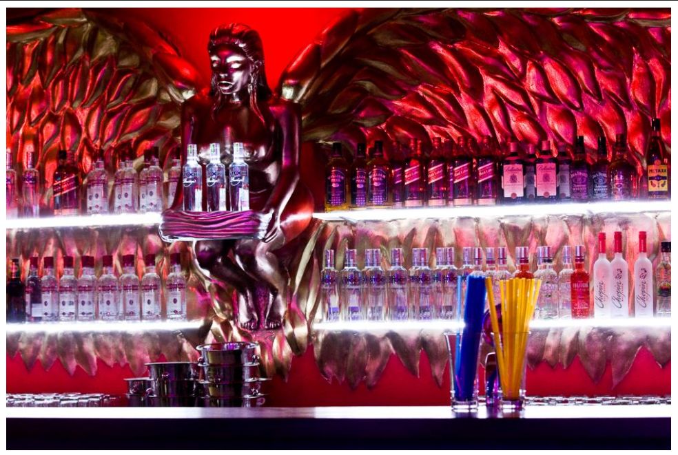 Bar w dyskotece z rzeźbą anioła i podświetlonymi półkami pełnymi butelek alkoholu, czerwone światło