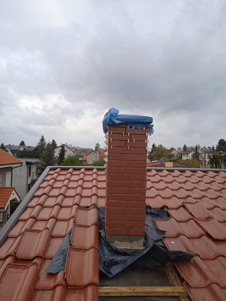 Murowany komin z cegły, zabezpieczony folią i plandeką na dachu z czerwonej dachówki, w tle zabudowa jednorodzinna i pochmurne niebo.