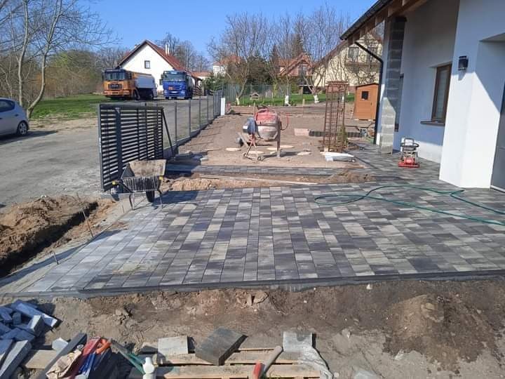 Układanie szaro-czarnej kostki brukowej na posesji, widoczny fragment ogrodzenia, betoniarka i narzędzia budowlane, w tle dom i dwa samochody ciężarowe.