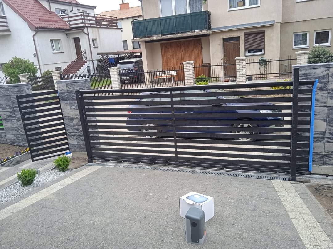 Nowoczesna, pozioma, przesuwna brama wjazdowa z czarnych profili, z automatyką na kostce brukowej. W tle widoczny dom jednorodzinny i zaparkowany samochód.
