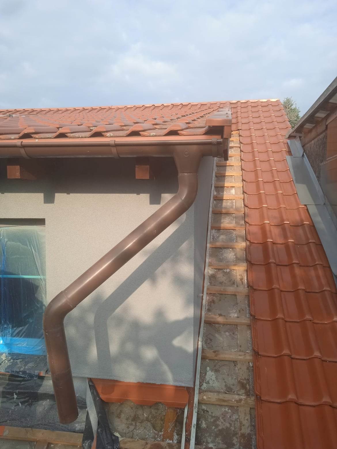Fragment elewacji budynku z brązową rynną, widoczne tynkowanie ściany i fragment dachu z czerwoną dachówką podczas prac wykończeniowych.