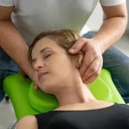 Kobieta z zamkniętymi oczami leży na zielonym fotelu, podczas gdy terapeuta masuje jej okolice szyi i głowy.