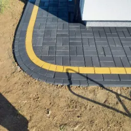 Ułożona, czarno-szara kostka brukowa z żółtym obramowaniem na zakręcie, widoczna butelka wody i rynna spustowa przy białej ścianie budynku.