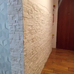 Wykończona ściana z dekoracyjnej białej cegły, sąsiadująca z trójwymiarową białą płytą ścienną i drewnopodobną podłogą, widoczne wystające kable elektryczne.