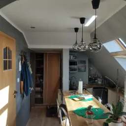 Jasne wnętrze poddasza z drewnianym stołem barowym, trzema lampami wiszącymi z ozdobnymi kloszami i widocznym skosem dachu z oknem.