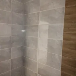 Ściana w łazience wyłożona szarymi, prostokątnymi płytkami imitującymi kamień, z pasem płytek imitujących drewno po prawej stronie. Widoczny narożnik pomieszczenia i błyszcząca powierzchnia płytek...