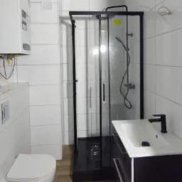 Wykończona łazienka z białą armaturą, czarną kabiną prysznicową i umywalką z szafką, białe płytki na ścianach, drewnopodobne płytki na podłodze, bojler elektryczny na ścianie.