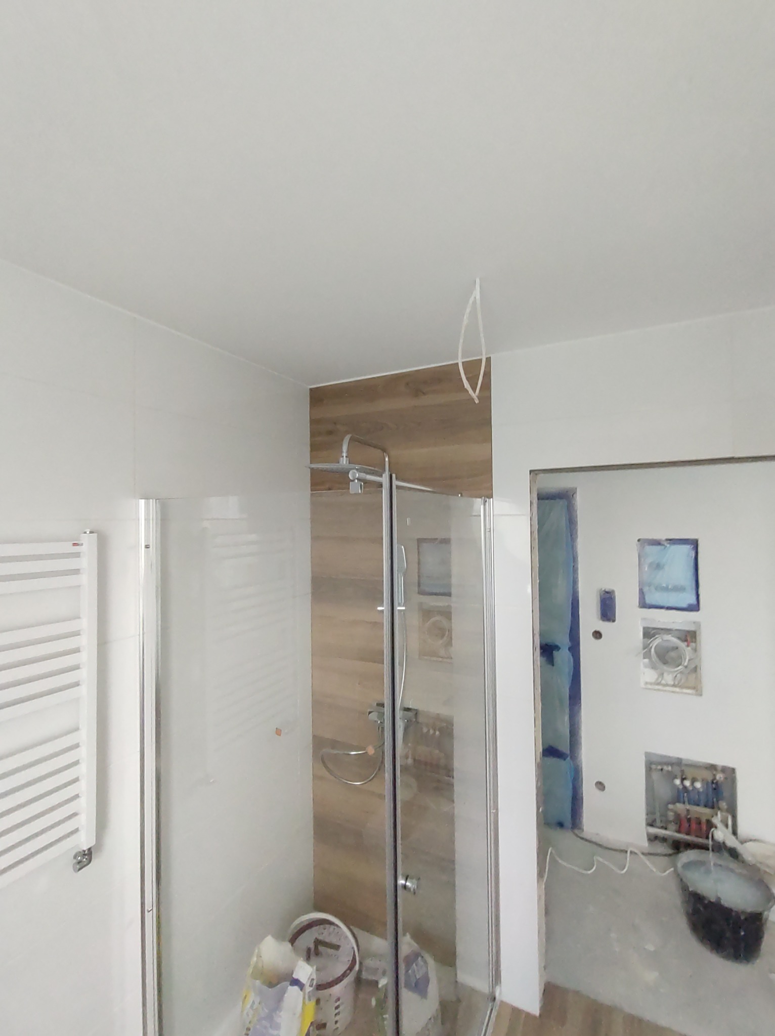 Wnętrze łazienki w trakcie remontu: kabina prysznicowa ze szklanymi drzwiami, grzejnik drabinkowy na ścianie, fragment ściany wyłożony płytkami imitującymi drewno, wiadro z zaprawą w tle, widoczne...