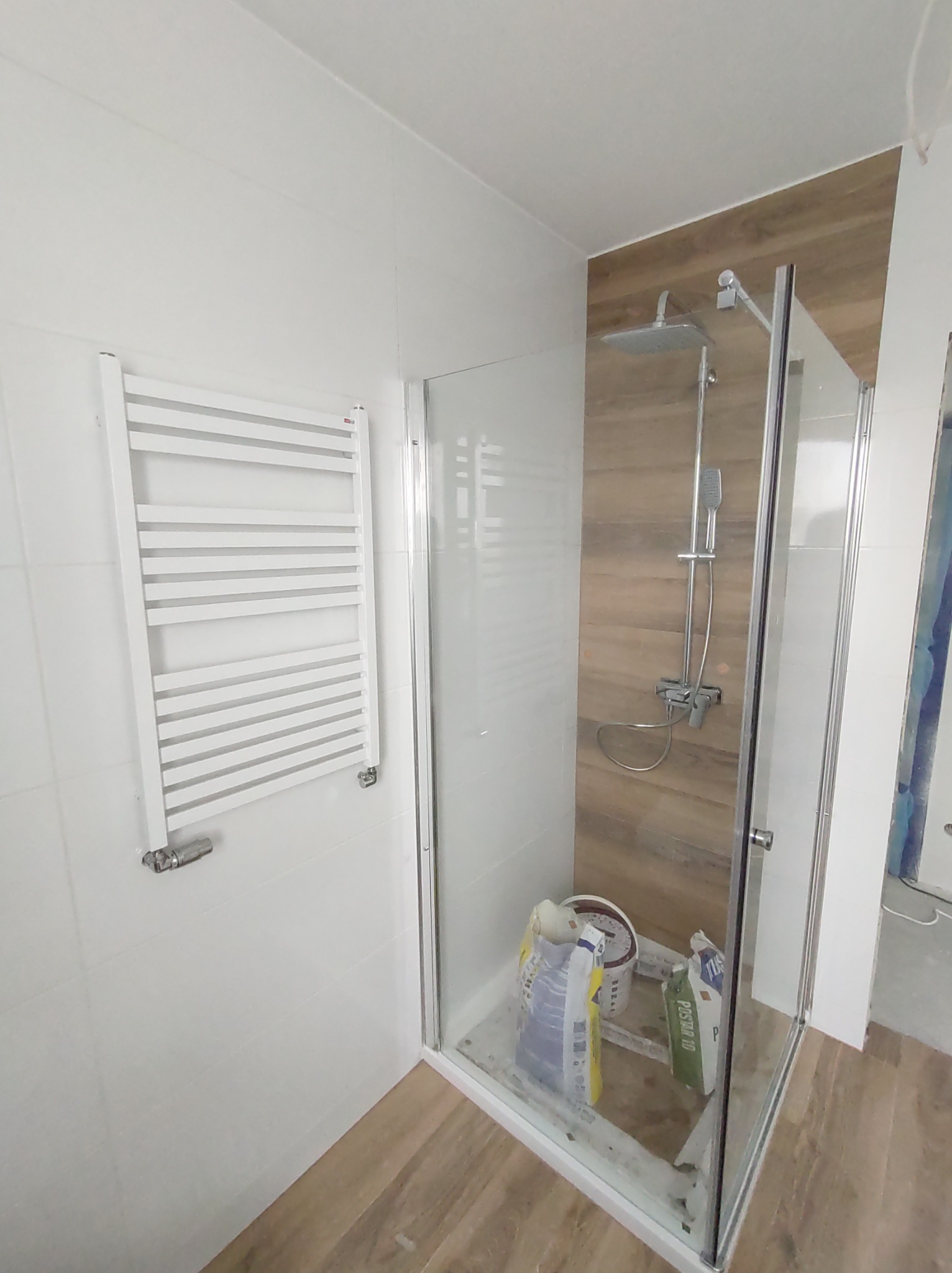 Remont łazienki w trakcie: kabina prysznicowa z deszczownicą na ścianie wyłożonej panelami imitującymi drewno, biały grzejnik drabinkowy na ścianie z białych płytek, widoczne worki z zaprawą...