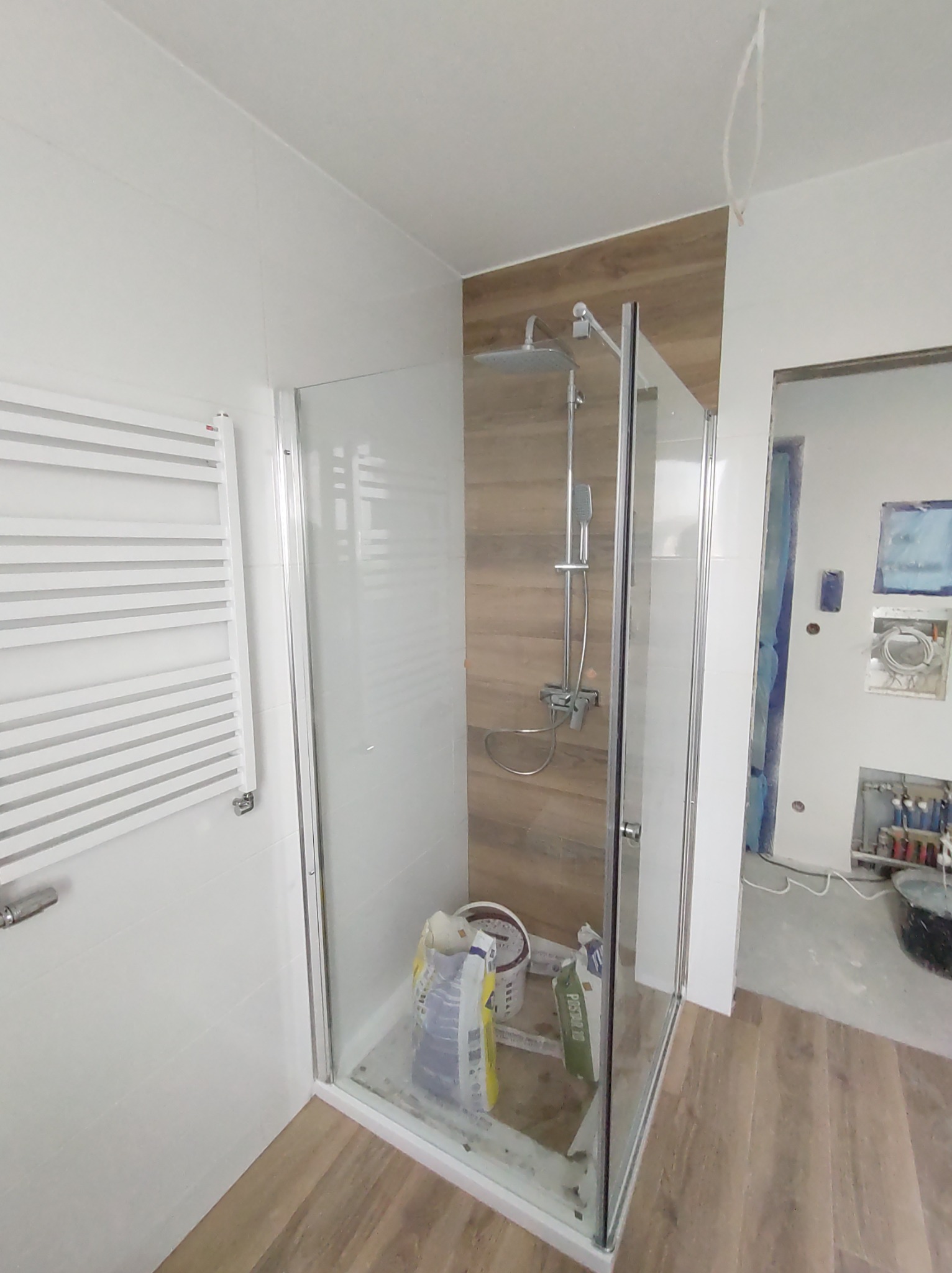 Szklana kabina prysznicowa z deszczownicą, częściowo wykończona drewnopodobną okładziną, widoczne białe płytki, grzejnik drabinkowy, worki z zaprawą i narzędzia wewnątrz kabiny, stan surowy...