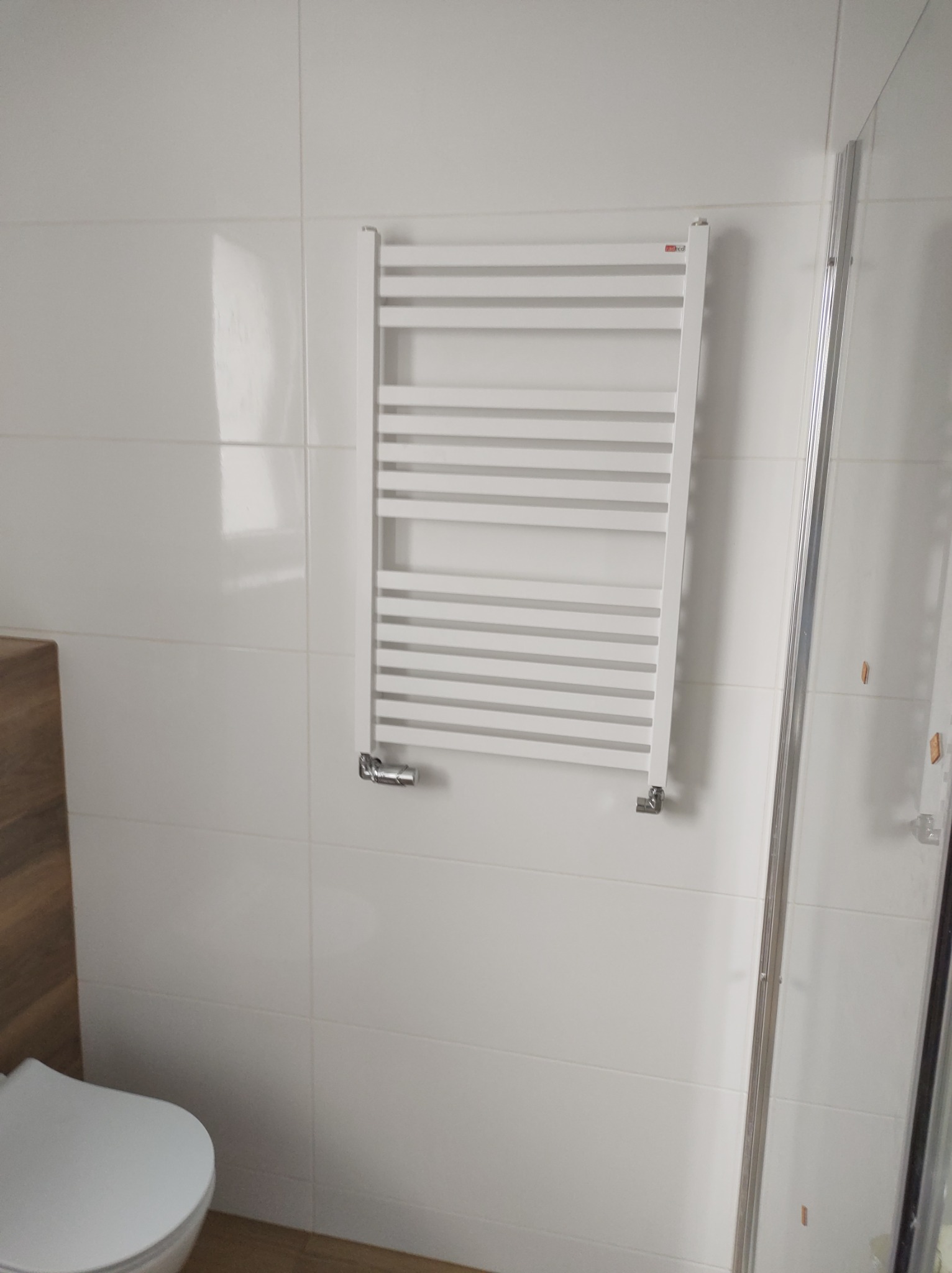 Biały grzejnik łazienkowy drabinkowy zamontowany na białych, błyszczących płytkach, widoczny fragment kabiny prysznicowej i toalety.