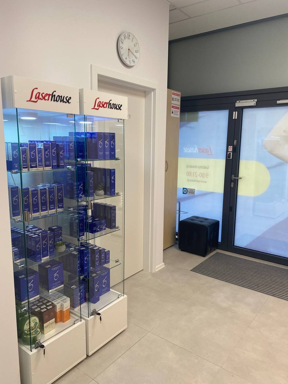 Wnętrze kliniki: witryna z kosmetykami marki Laserhouse, zegar na ścianie, drzwi wejściowe z widocznym napisem Laserhouse odbitym w szybie.