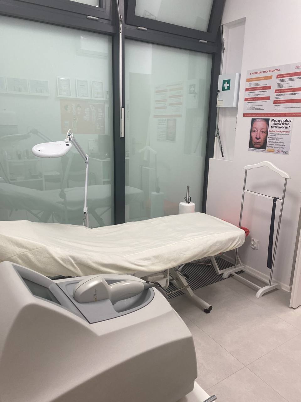Wnętrze gabinetu z białym leżanką kosmetyczną, lampą powiększającą i urządzeniem do zabiegów na ciało; na ścianie apteczka pierwszej pomocy i plakat informacyjny.
