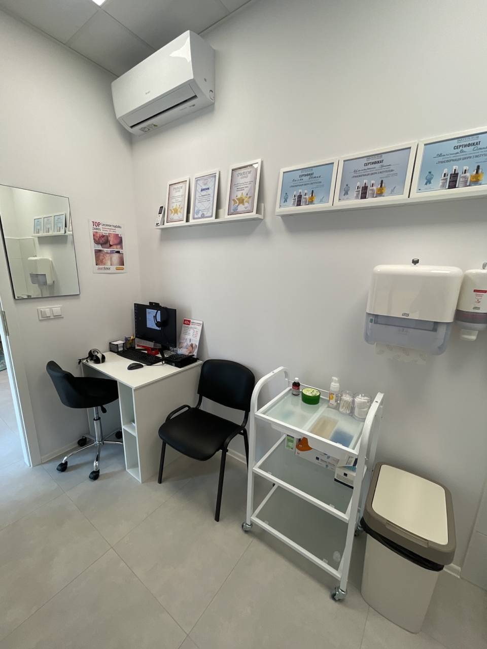 Jasne wnętrze gabinetu z biurkiem, krzesłami, wózkiem z akcesoriami i certyfikatami na ścianie.