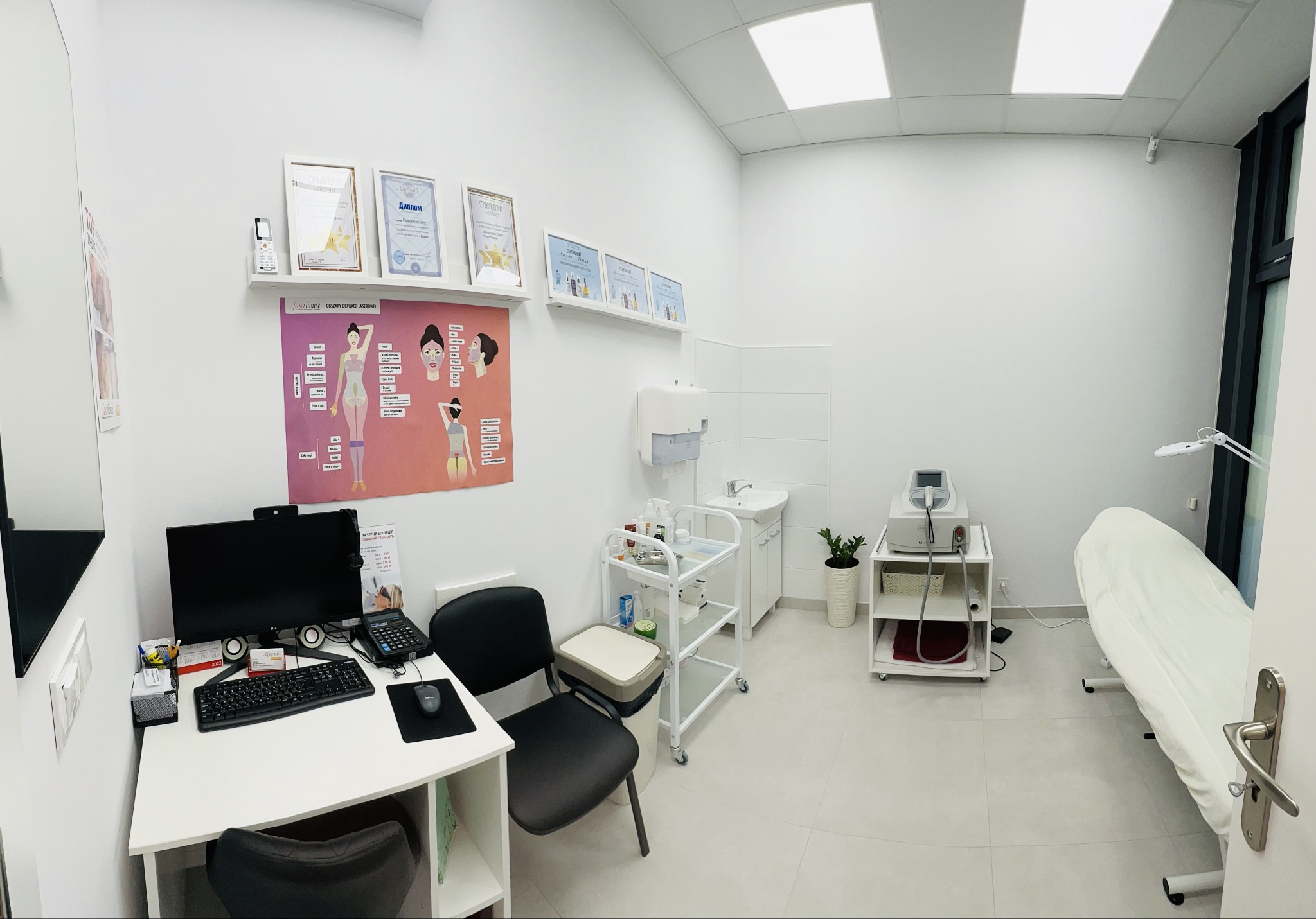 Jasne, sterylne wnętrze gabinetu medycyny estetycznej z biurkiem, komputerem, wózkiem z kosmetykami, aparaturą zabiegową i leżanką przykrytą białym prześcieradłem, na ścianach dyplomy i plansze...