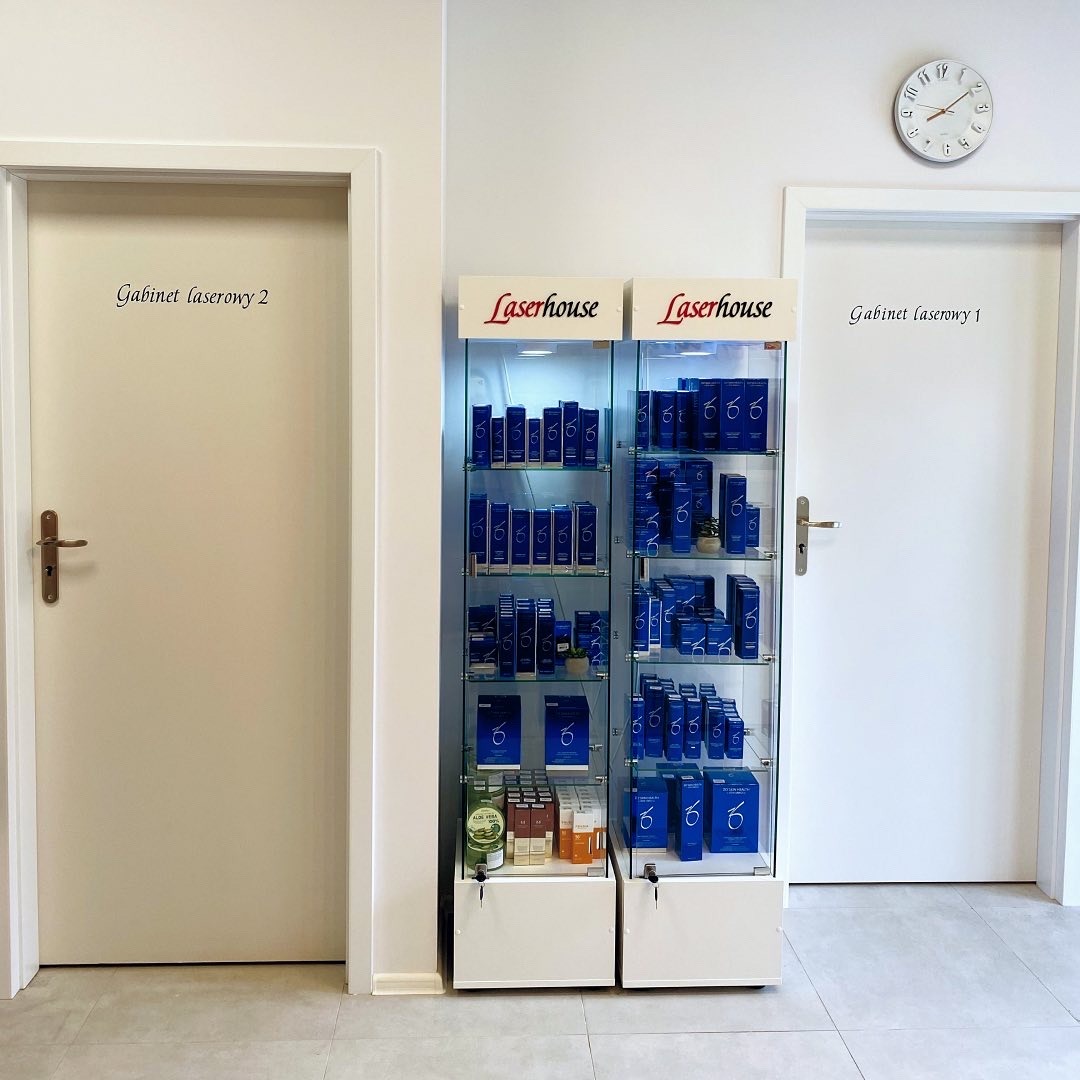 Wnętrze gabinetu z witryną wypełnioną niebieskimi opakowaniami kosmetyków marki Laserhouse, widoczne drzwi do gabinetów laserowych o numerach 1 i 2, zegar na ścianie.