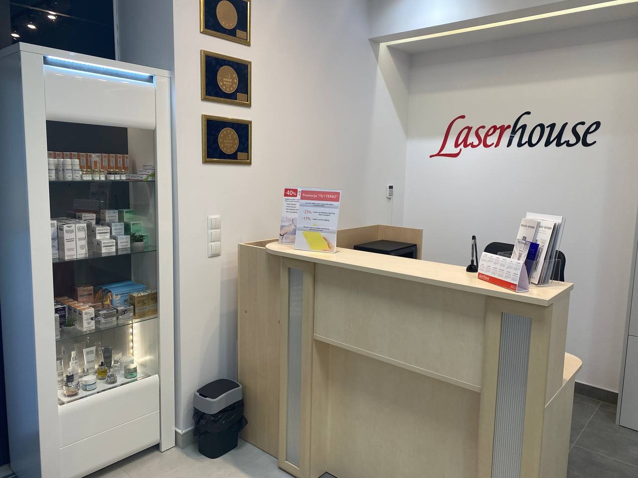 Jasne wnętrze kliniki z recepcją, witryną z kosmetykami i logo 'Laserhouse' na ścianie, wiszące dyplomy w złotych ramach.