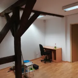 Pokój z drewnianą konstrukcją stropu, biurkiem, krzesłem i podłogą z paneli, widoczne narzędzia malarskie i wiadro z farbą, sugerujące przygotowania do prac remontowych.