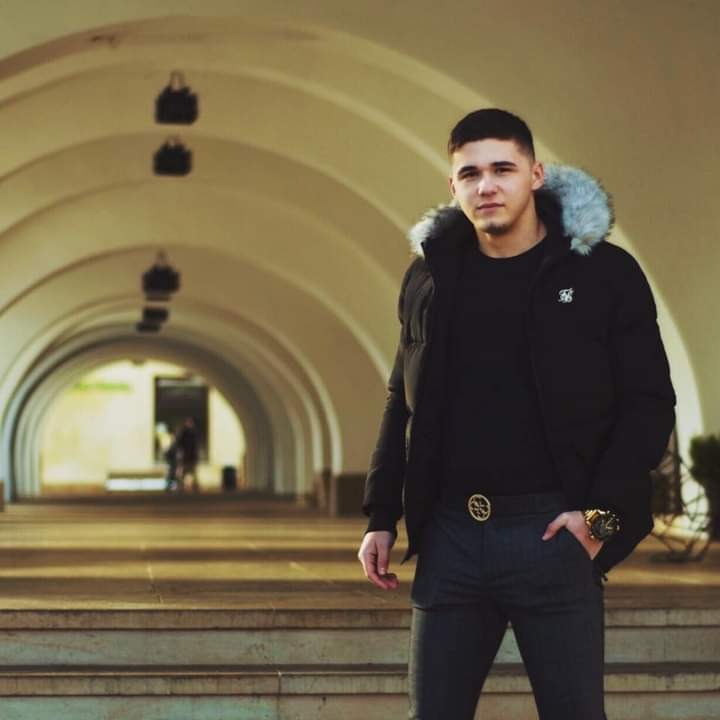 Młody mężczyzna w czarnej kurtce z futrzanym kapturem, pozujący na tle architektonicznej konstrukcji z łukami.