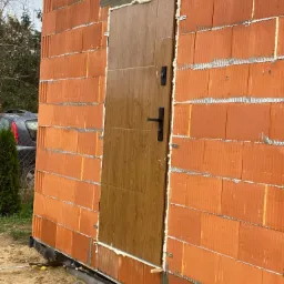 Montaż brązowych drzwi zewnętrznych z czarną klamką w surowym murze z pustaków ceramicznych, widoczne pianowanie uszczelniające, tło z zielenią i fragmentem samochodu.