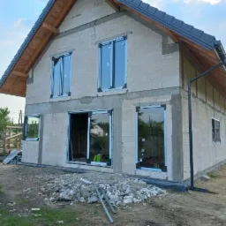 Surowy stan domu w trakcie budowy z zamontowanymi oknami i drzwiami w czarnych ramach, widoczne materiały budowlane i rury na ziemi.