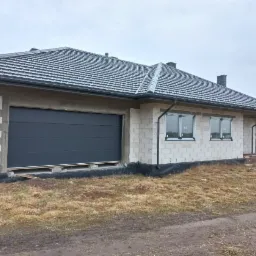 Szara brama garażowa segmentowa w surowym stanie budynku z białego bloczka, widoczne okna i dach z szarej dachówki, krajobraz polny w tle.