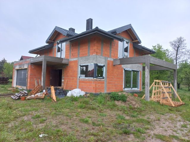 Dom jednorodzinny w trakcie budowy z zamontowanymi oknami z czarnymi ramami, widoczne pustaki i elementy konstrukcyjne, na pierwszym planie teren budowy z paletami i materiałami budowlanymi.