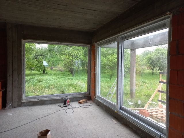 Wnętrze surowego domu z dużymi oknami przesuwnymi z widokiem na zieleń, widoczne narzędzia i drabina, etap montażu.